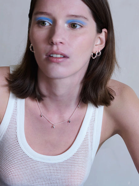 Woman with blue eyeliner wearing a white tank top with arrow spike silver necklace against a plain background