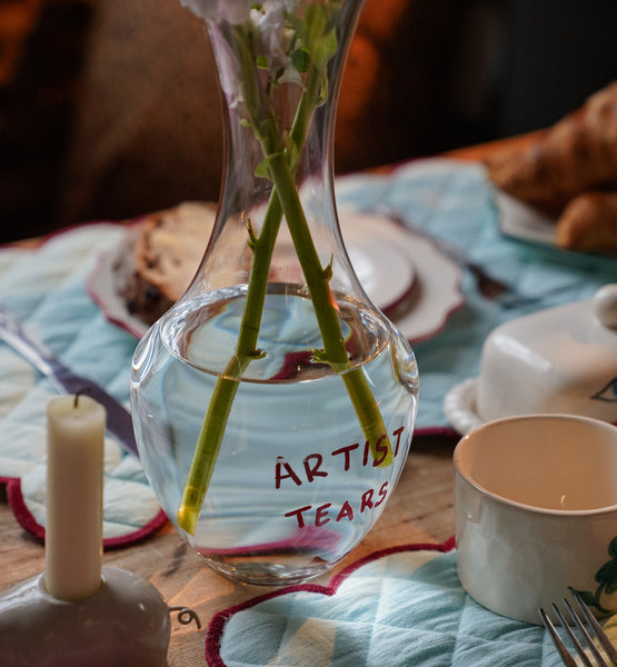 Clear glass vase with 'Artist Tears' text on a table setting with bread and a candle.