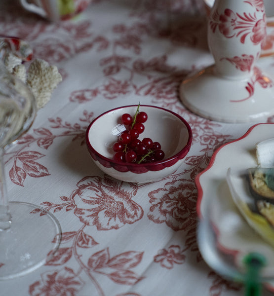 Small ceramic bowl with painted red scalloped edge with red berries in it on floral-patterned tablecloth with decorative items.