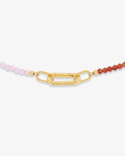 A close-up of a beaded necklace with a gold chain-link clasp engraved with 'RACHEL JACKSON'. The left side of the beads is light pink, and the right side is orange-red, set against a clean white background.