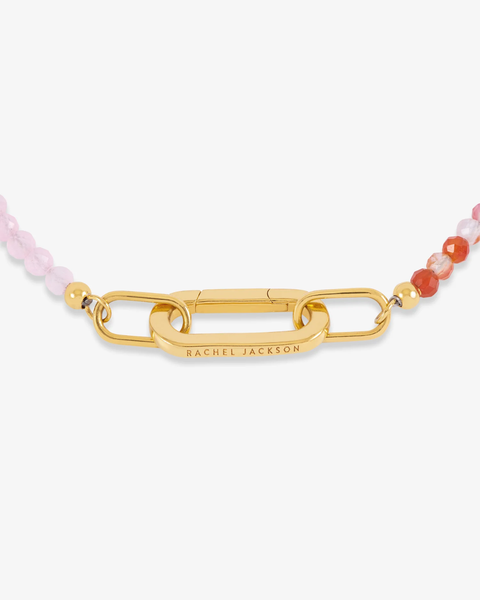 A close-up of a gold-tone bracelet with a central rectangular link engraved with 'RACHEL JACKSON'. The chain is flanked by small pink and orange faceted beads on a clean white background.