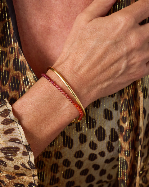 A close-up of a person's wrist wearing two bracelets: a polished gold bangle and a thin red beaded bracelet, against a leopard-print garment.
