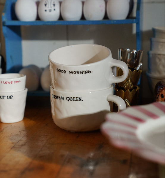Two white cappuccino mugs with text on a wooden surface, with a shelf of ceramic items in the background.