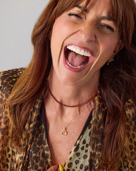 A close-up shot of a woman with long brown hair, wearing a leopard print top and layered necklaces, captured in a moment of laughter against a plain light gray background.