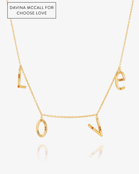 A gold-tone necklace spelling out the word LOVE, with each letter hanging from a short chain segment. The necklace is displayed on a clean white background with a subtle reflection below.