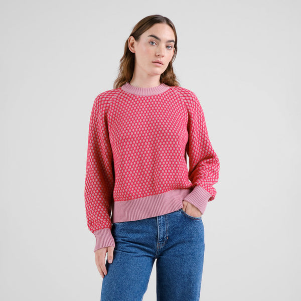 Woman wearing a red and pink tight bubble knit jumper and blue jeans on a light gray background