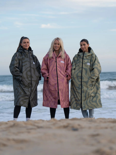 Three people wearing long, patterned camo changing robes on a beach.
