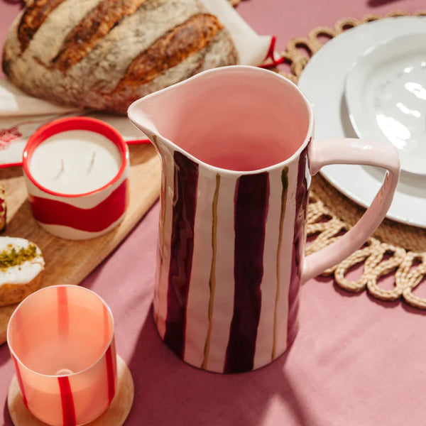 Pink and maroon striped jug on a table with bread, plates, and candles