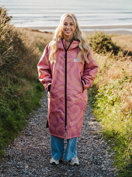 Person wearing a pink camo changing robe standing on a path near the sea.