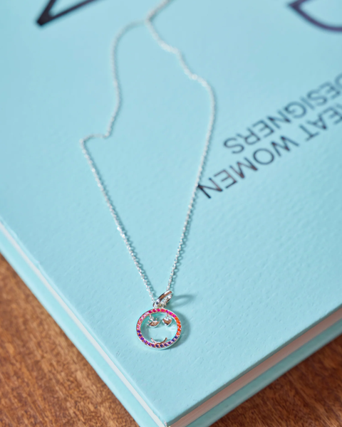 A silver chain necklace with a small circular pendant featuring a colorful rainbow border and a central happy face design, resting on a light blue book over a wooden surface.