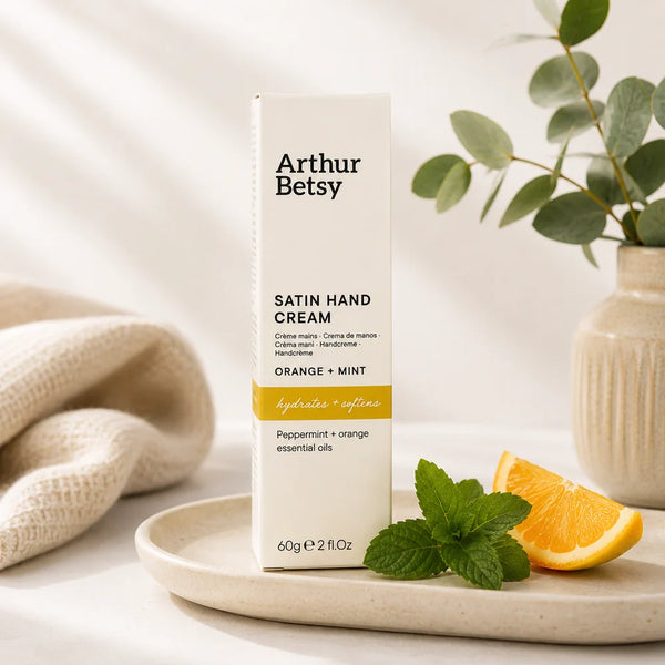 Arthur Betsy Satin Hand Cream in Orange + Mint scent, shown in a tall white box with black text and a yellow-orange band, styled on a ceramic tray with mint leaves, an orange slice, and a neutral knit blanket.