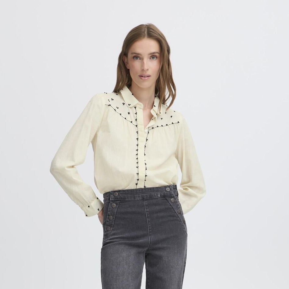 Woman wearing a beige blouse with black embroidery and dark jeans on a white background