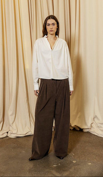 Person wearing a white blouse and brown pants standing in front of beige curtains.