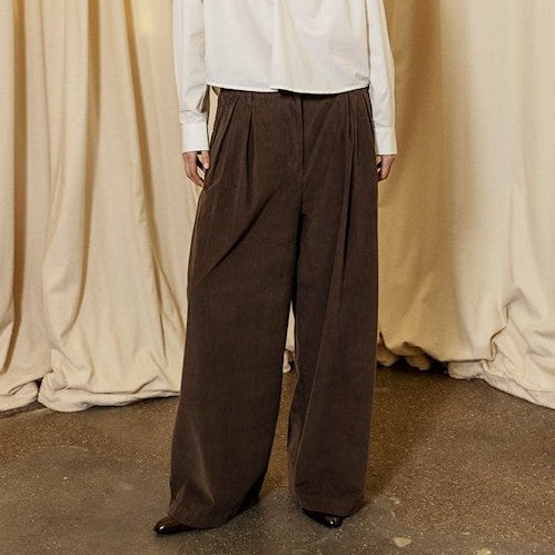 Person wearing a white shirt and brown wide-leg pants standing against a beige curtain backdrop.