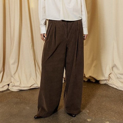 Person wearing a white shirt and brown wide-leg pants standing against a beige curtain backdrop.