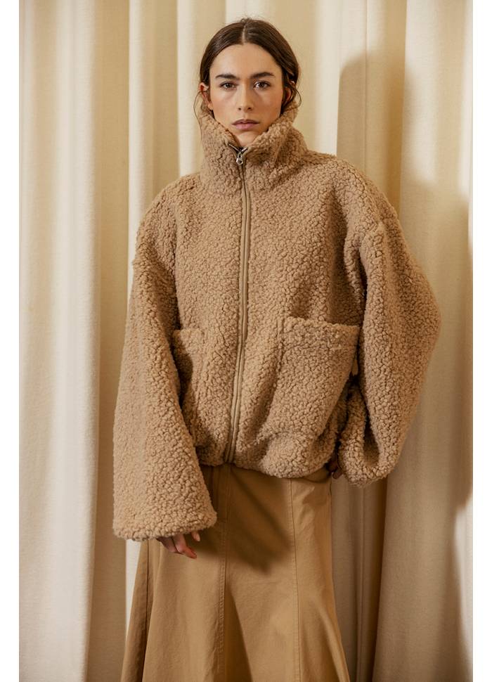 Person wearing a beige fleece jacket and pants against a beige curtain background