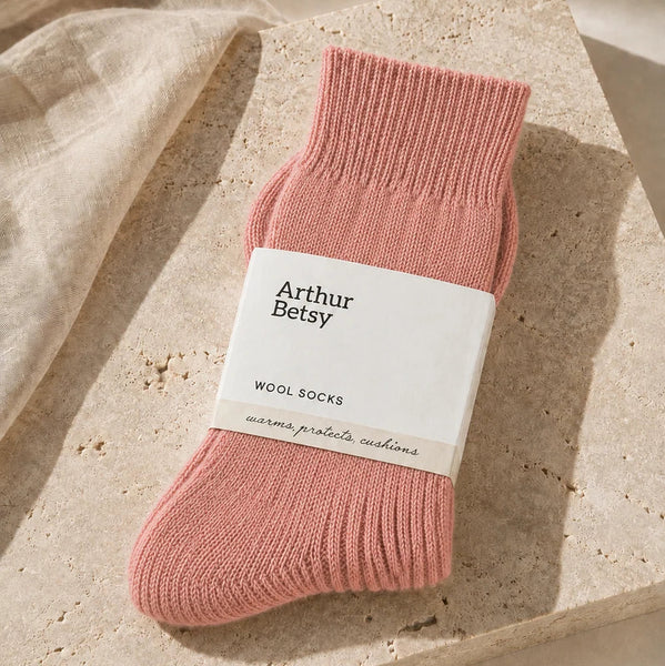 A single pink knit sock with a white paper band reading 'Arthur Betsy WOOL SOCKS' is styled on a beige stone slab with soft linen fabric in the background.